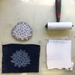 A flat-lay of a handmade block print and ink-making setup on a light beige surface. The set includes a carved floral mandala wood block, a dark indigo denim fabric square with the matching floral print, ink  brayer, and a white ink swatch rolled out in a square shape.