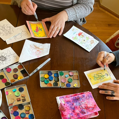 Two people sitting at a table with paints, watercolor brushes, and painting botanical themed images.