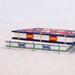 Two decorative handmade paper notebooks stacked on a light surface. The top notebook has a navy blue cover with a red and white floral pattern and yellow cord binding.