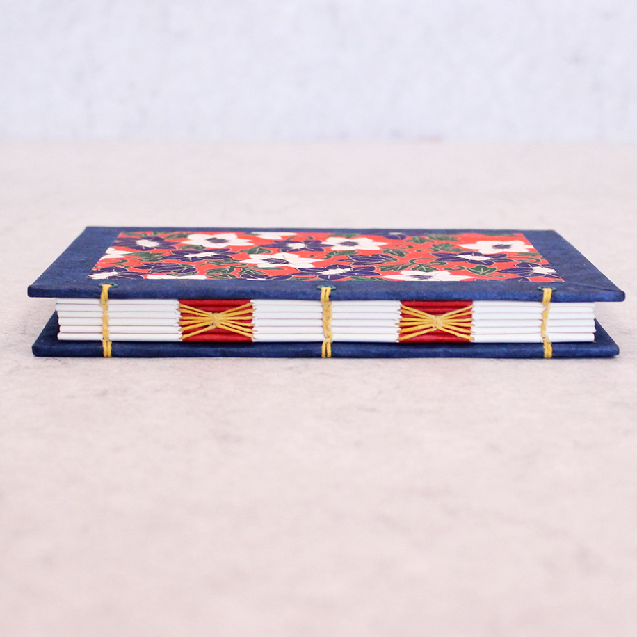 A small decorative book with a navy blue cover, a colorful floral pattern on the top, white pages, and gold thread binding, photographed on a light beige surface against a pale gray background.