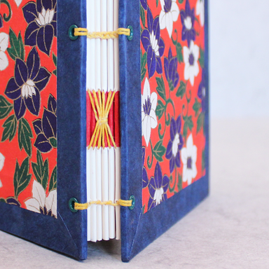 A close-up of a handmade paper book with a blue cloth spine and floral-patterned covers in red, navy, white, and green, bound with yellow thread.