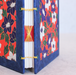 A close-up of a handmade paper book with a blue cloth spine and floral-patterned covers in red, navy, white, and green, bound with yellow thread.
