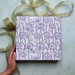 Handmade Indian Cotton Paper- Block Printed lotus floral pattern with lavender flowers and green stems used as gift wrap. Shown with a hand and green ribbon.