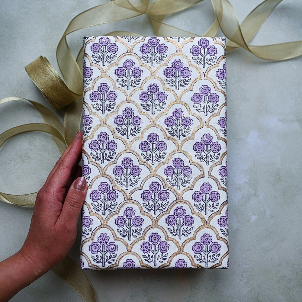 Block Printed lavender and green floral pattern with gold arches used to wrap a gift. Shown with a hand and gold ribbon.