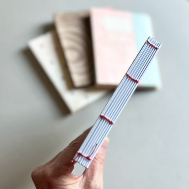 A hand holds a sewn text block of pages stitched with thin red thread, while three soft-focus notebook covers sit on a light surface in the background.
