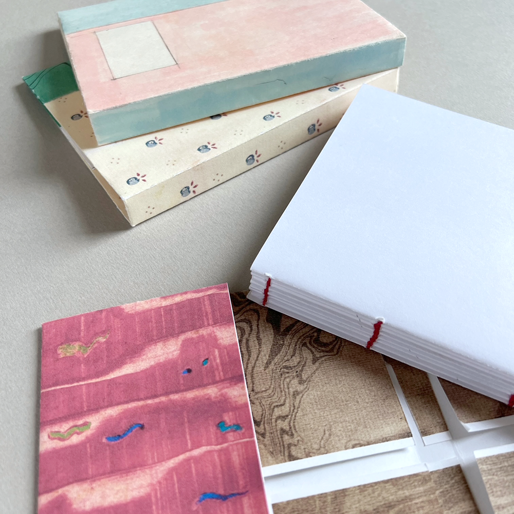 Handmade paper notebooks on a neutral beige surface. The stack shows three distinct covers: a top one with a textured pink and light blue design, a middle one with a cream base and small blue floral motifs, and a bottom one showing a pink textured cover with blue and gold abstract details with a white text block and red stitching