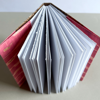 An open handmade accordion-style journal with white pages fanned out, bound with a pink patterned cover and a brown patterned spine, photographed on a neutral beige surface.