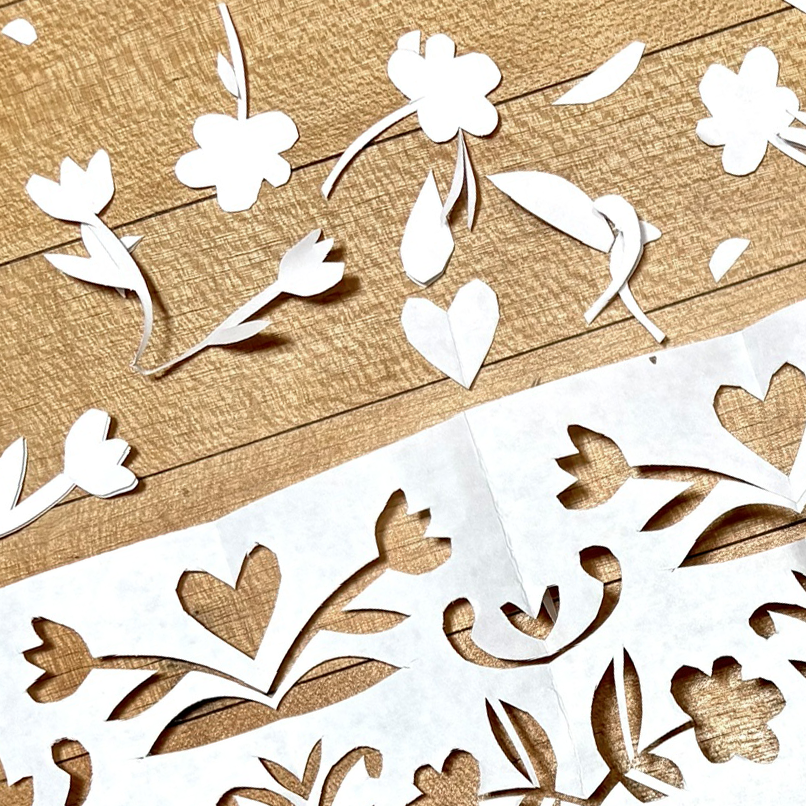 White floral paper cuts laid out on a textured brown wood background, showing cutouts of flowers, leaves, and hearts