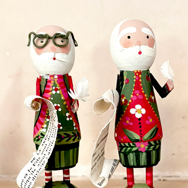 Two hand-painted wooden figurines of Santa Claus with glasses and a long paper list, standing against a plain beige background.