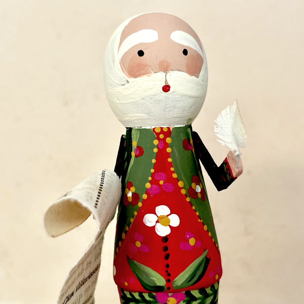 A handcrafted wooden Santa Claus doll with a white paper beard and hat, holding a rolled paper list, dressed in a red and green patterned coat with a white flower on the front, set against a plain beige background.