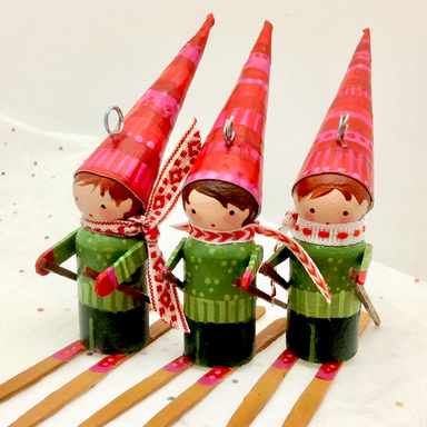 Three small skier ornaments wearing red patterned pointed hats and green striped outfits, posed on wooden sticks against a plain light background.