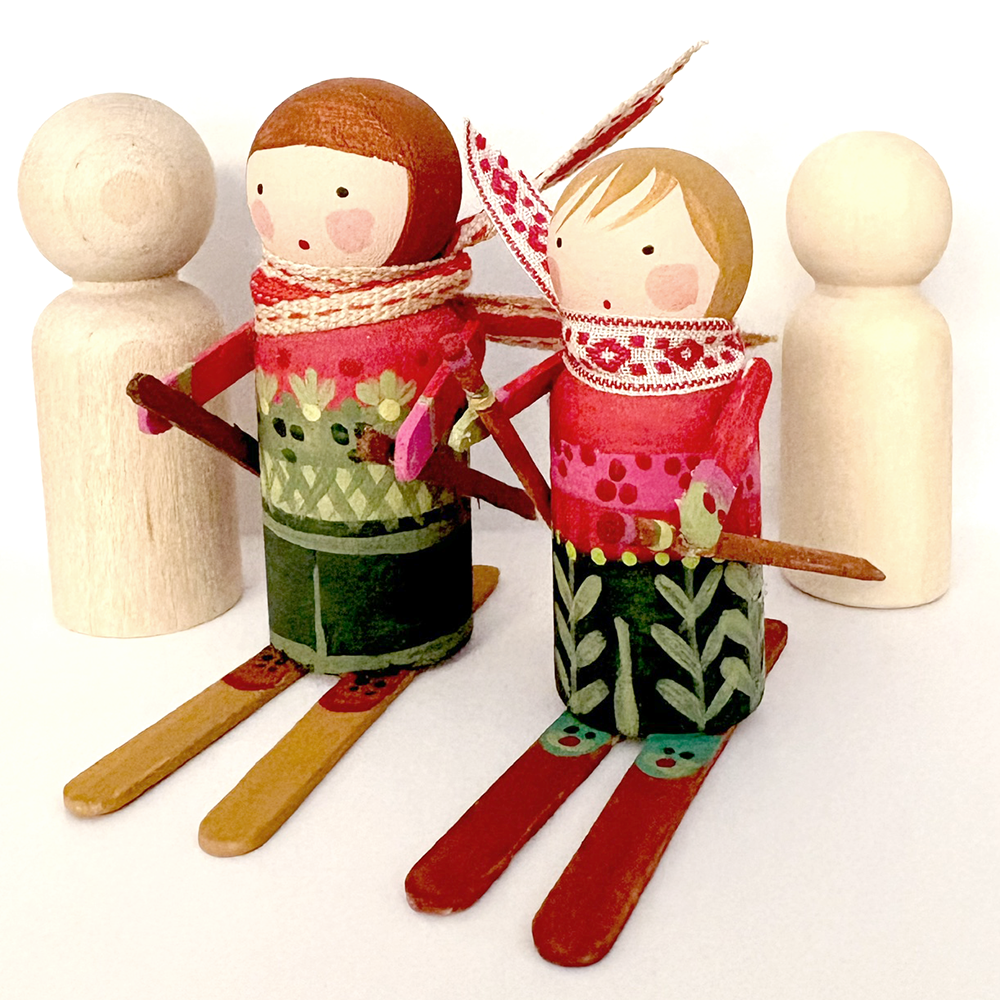 Handcrafted wooden peg dolls dressed in winter clothing, with one doll wearing red and green patterned clothing and holding a ski pole, accompanied by two plain wooden peg dolls on a white background.