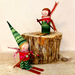 Two handmade wooden gnome skiers displayed on and around a rustic tree stump. One gnome is standing on skis in the foreground wearing a green patterned hat and red outfit, while the other sits on the stump in the background wearing a green sweater and red scarf.
