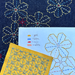 A close-up of a fabric craft project showing a dark indigo denim base with embroidered geometric and floral patterns in yellow, blue, and red. A light blue paper template with embroidered outlines and a handwritten color key are placed on top of the denim, alongside a yellow fabric swatch with blue geometric embroidery.