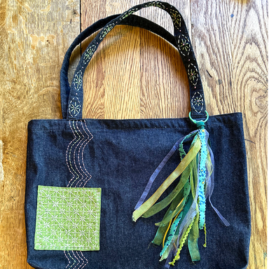 A handmade dark indigo denim tote bag with a green embroidered patch and a multi-colored ribbon tassel, displayed on a wooden surface.