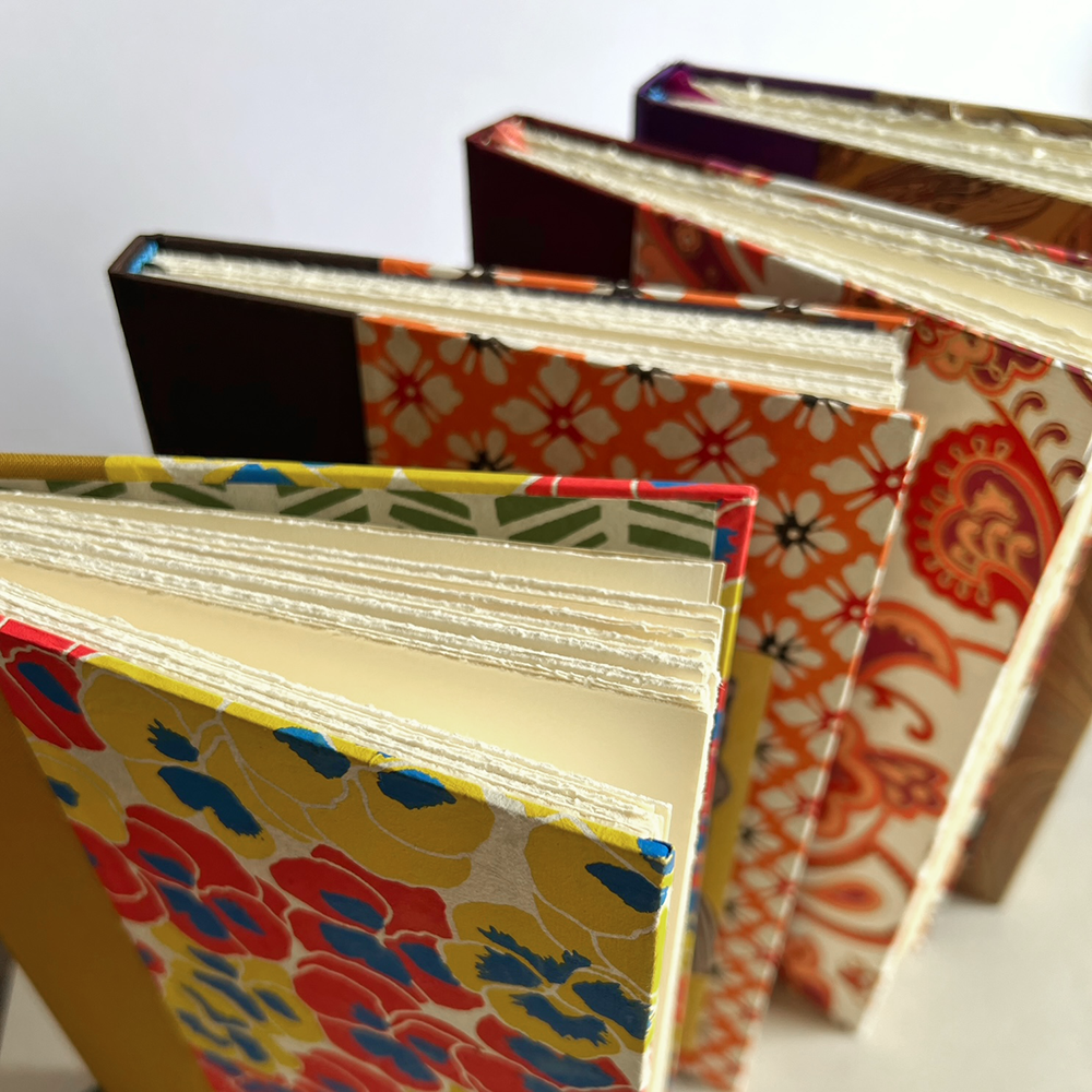 A close-up of several patterned journals standing upright, showing cream-colored pages and colorful covers with floral and paisley designs.