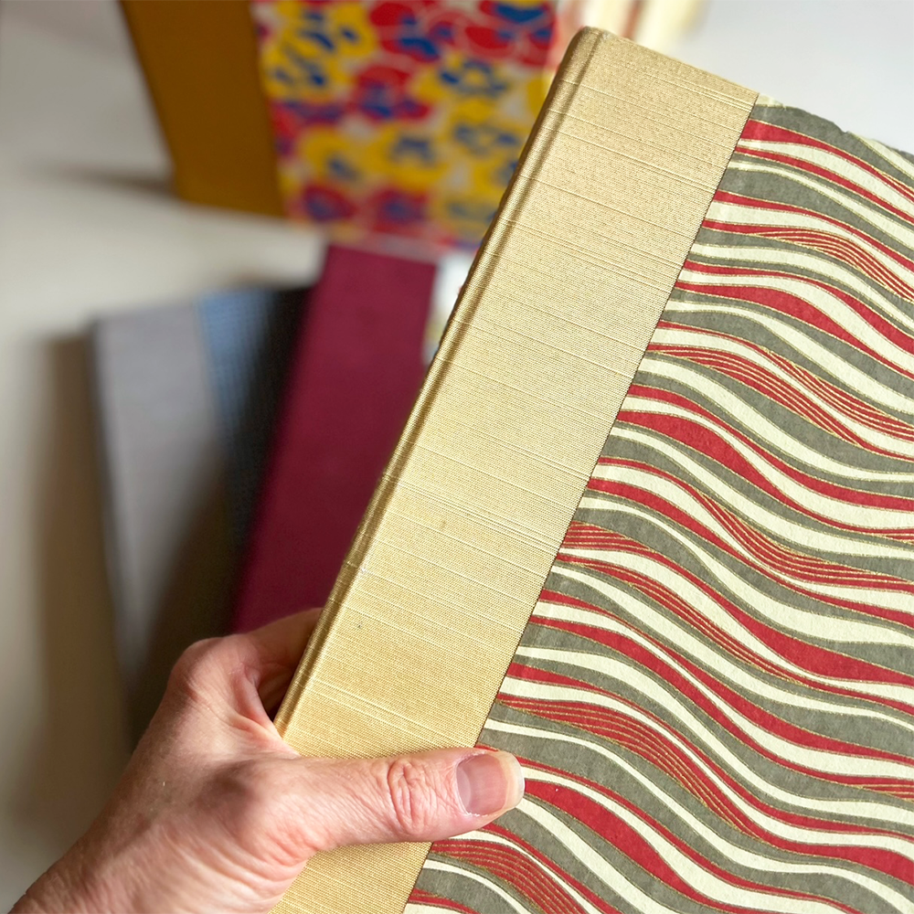 A hand holding a hardcover journal with a textured yellow cloth spine and a patterned cover featuring wavy red, olive green, and cream stripes with several books out of focus in the background.
