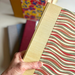 A hand holding a hardcover journal with a textured yellow cloth spine and a patterned cover featuring wavy red, olive green, and cream stripes with several books out of focus in the background.