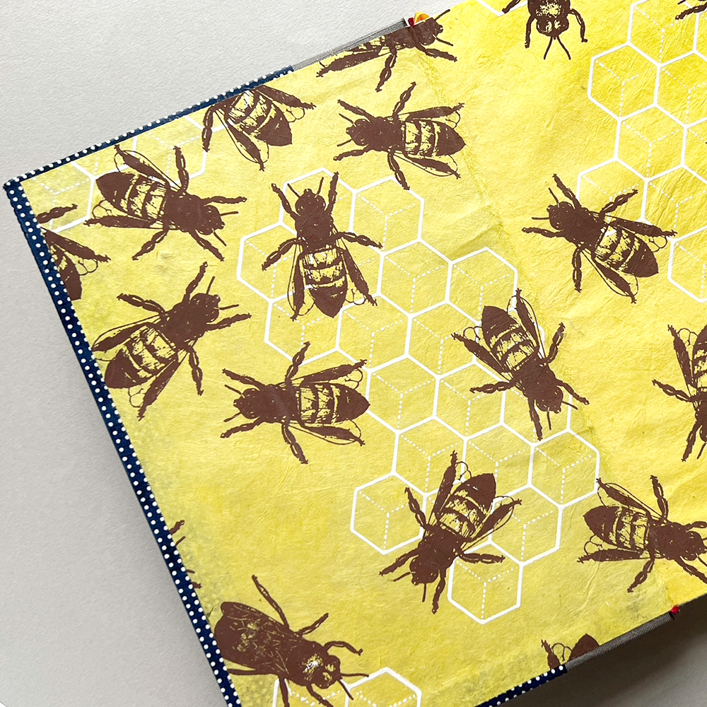 An open book featuring a yellow background with a pattern of brown and black bees and white hexagonal honeycomb cells, finished with a dark blue and white dotted border.