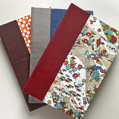 A stack of three hardcover journals fanned out on a light neutral background. The covers feature various patterns and colors,  a floral orange and white pattern, a gray linen-textured cover, a blue polka-dot cover, and  floral paisley pattern with red, blue, and green details.