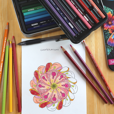 A flat-lay of colored pencils and drawing tools on a wooden surface, featuring a completed colorful word mandala drawing on white paper with the word 'COOPERATION' above it. An open metal pencil case, loose colored pencils, a pen, and part of a phone screen are visible.