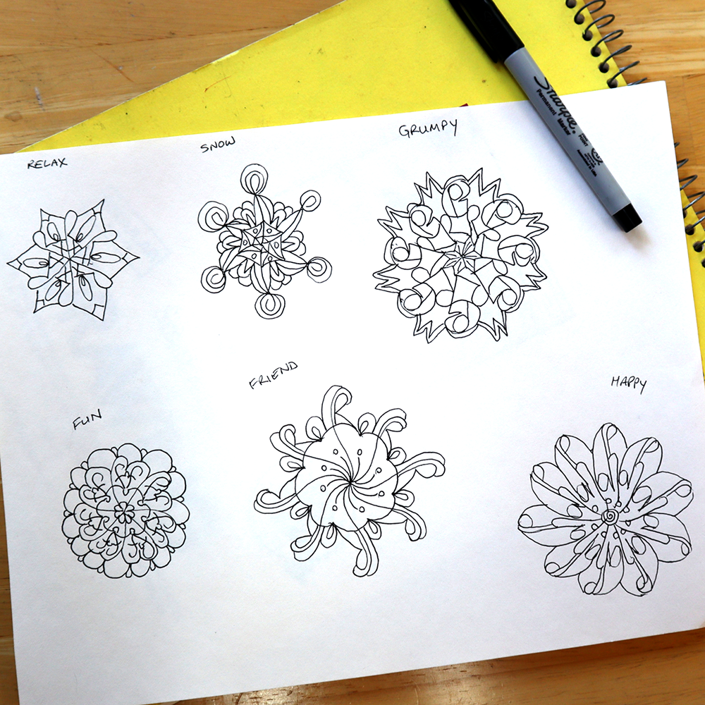 A sheet of white paper on a wooden table showing six black line-art mandala designs labeled Relax, Show, Grumpy, Fun, Friendly, and Happy. A yellow spiral notebook and a black Sharpie marker are partially visible around the paper.