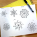 A sheet of white paper on a wooden table showing six black line-art mandala designs labeled Relax, Show, Grumpy, Fun, Friendly, and Happy. A yellow spiral notebook and a black Sharpie marker are partially visible around the paper.